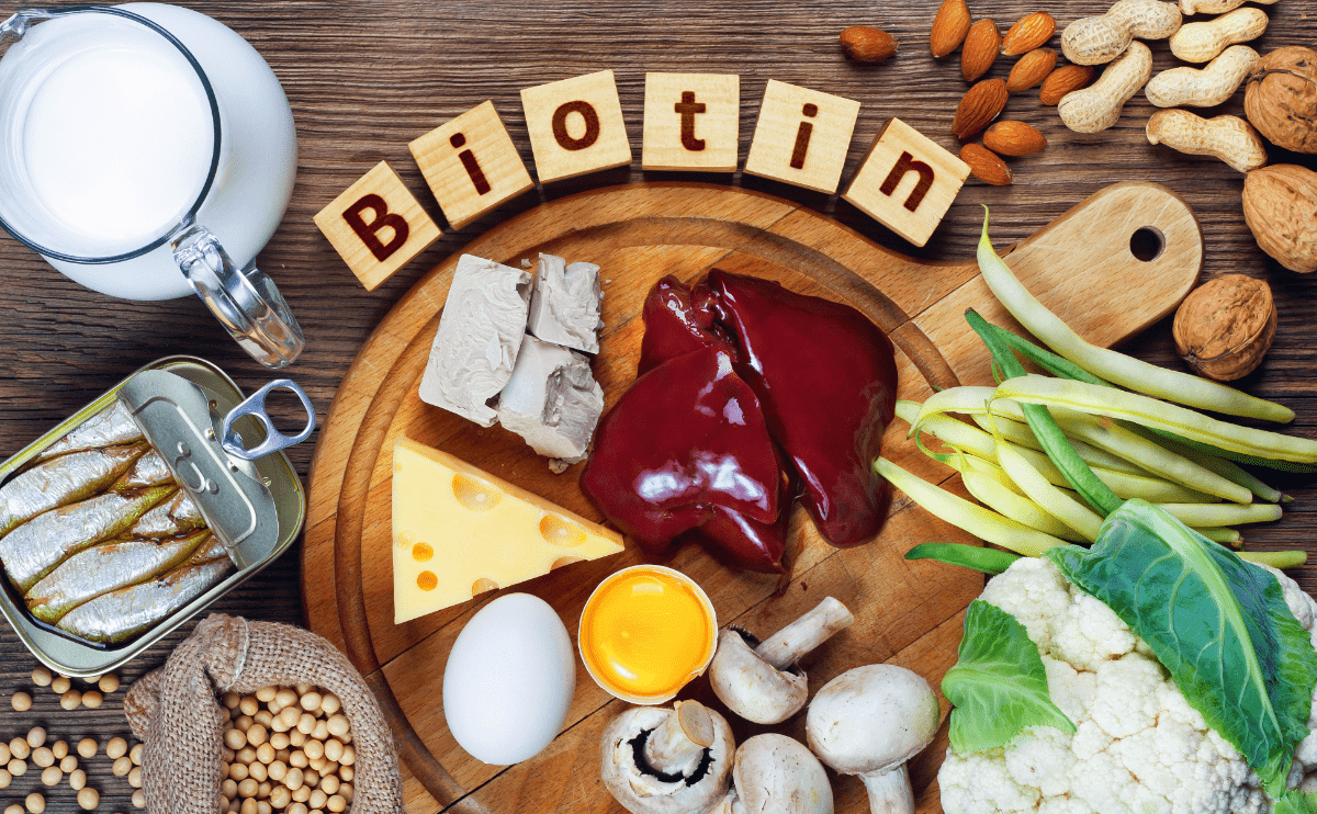 Įvairūs biotino šaltiniai, tokie kaip kepenys, sūris, kiaušiniai, žiediniai kopūstai, riešutai, sardinės ir grybai, padėti ant medinės lentelės – maisto produktai, kuriuose gausu biotino plaukų, odos ir nagų sveikatai.